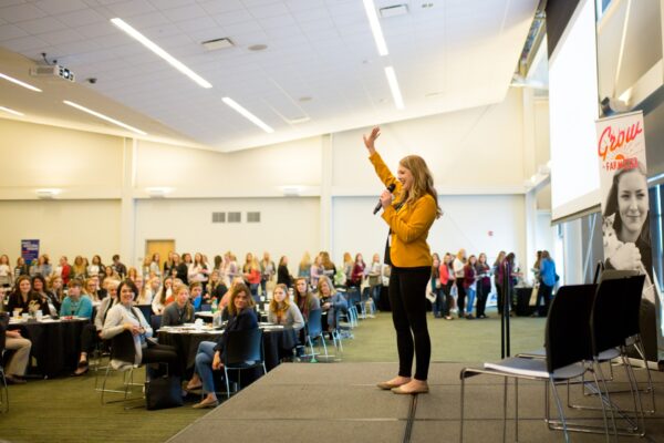 Photo of woman standing infront of an audience speaking with a microphone.
